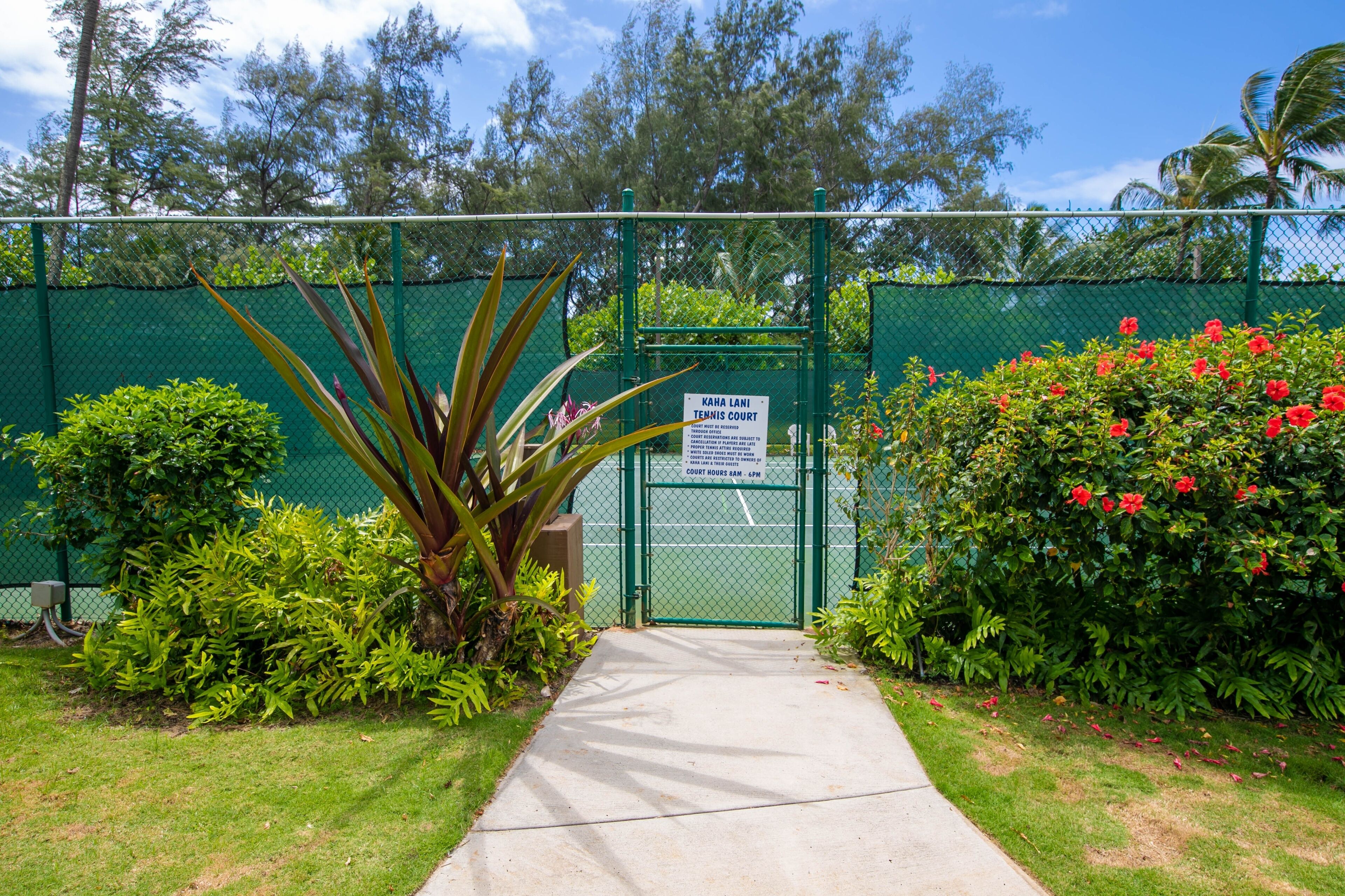 Entrada a una cancha de tenis con valla verde, un cartel con el horario y follaje circundante.