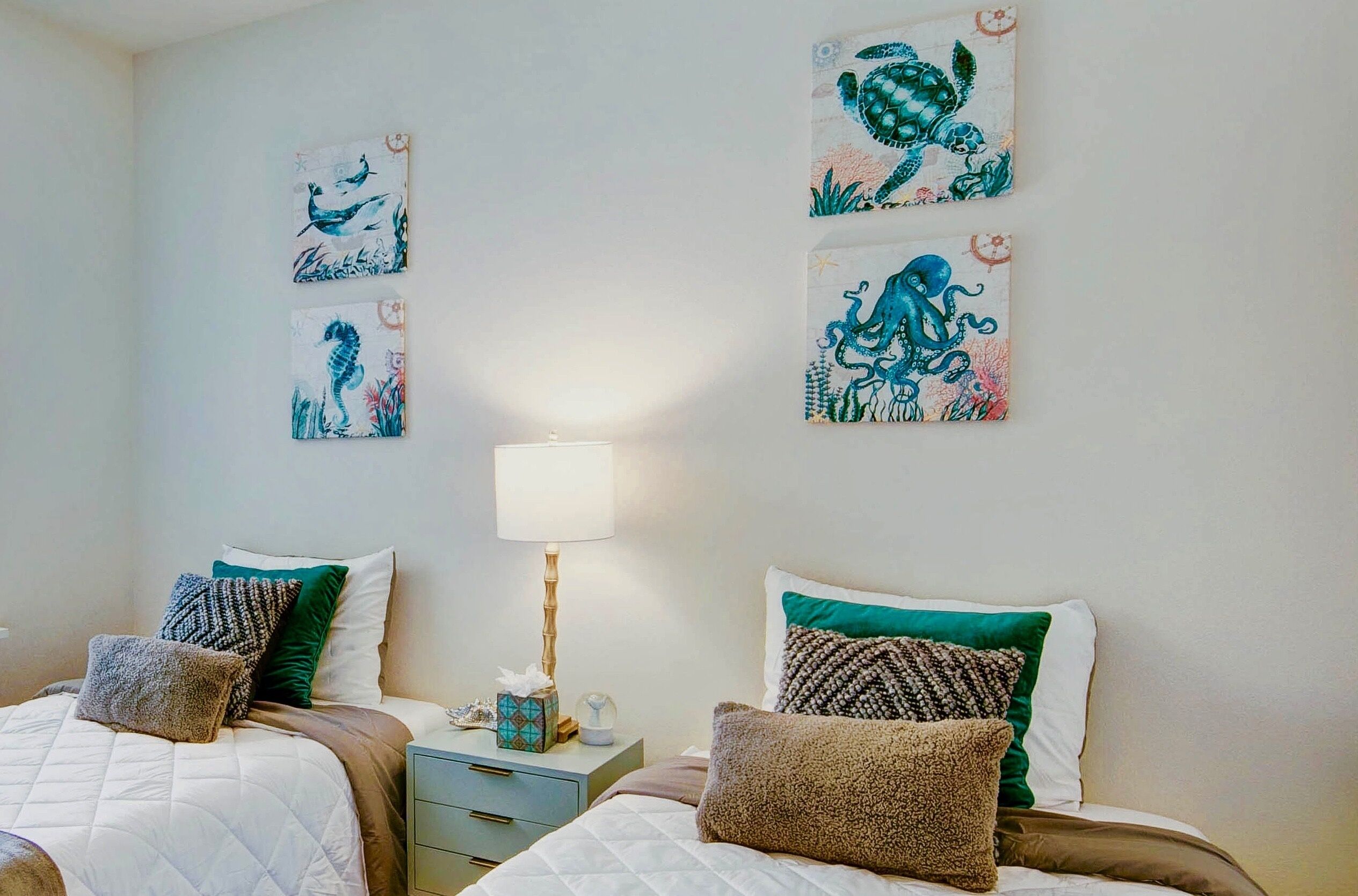 A bedroom with two beds, each with a quilted bedspread and multiple pillows. There is a bedside table with a lamp and a decorative item. The walls are adorned with three framed artworks depicting sea creatures.
