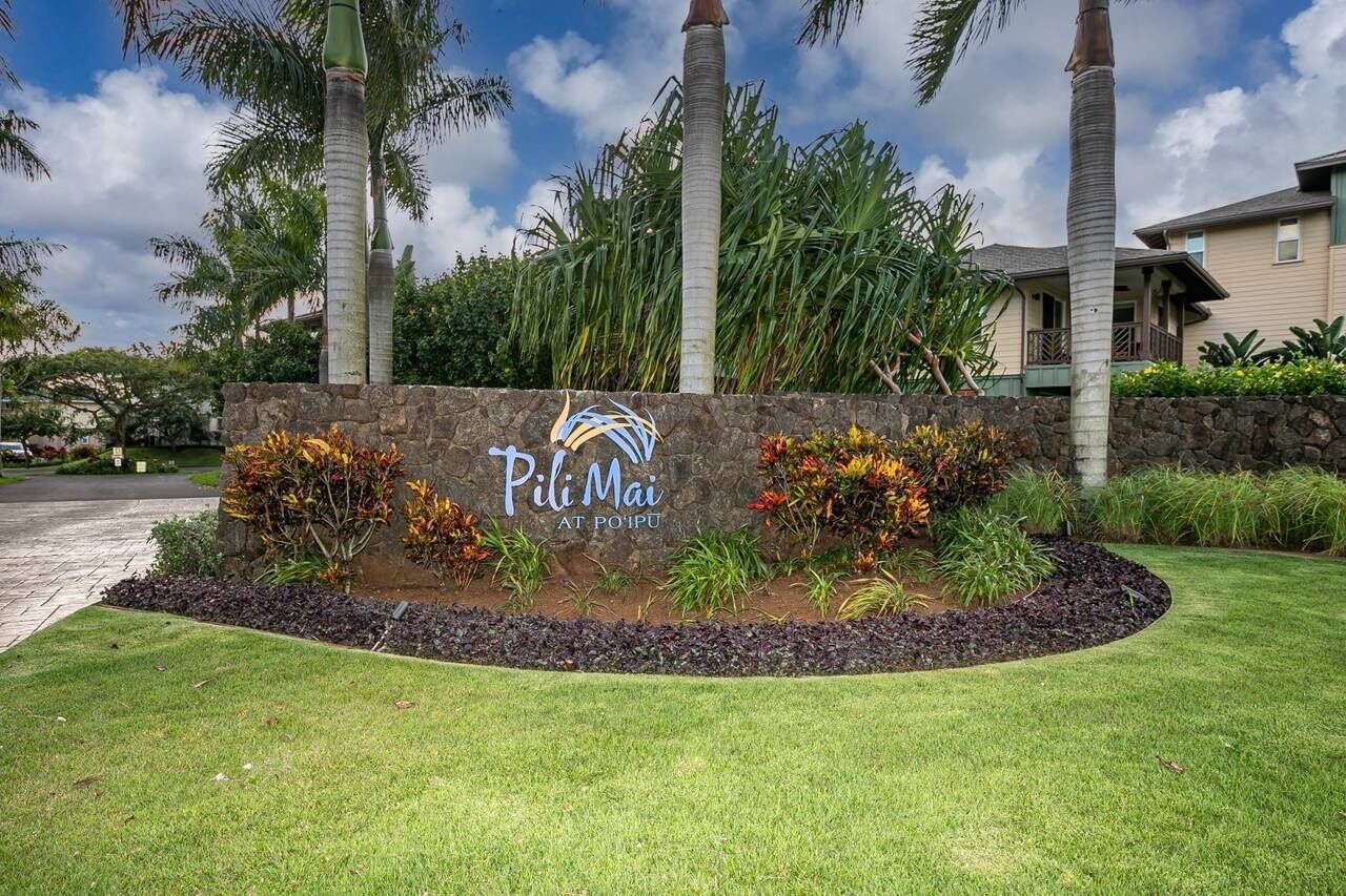 A sign for Pili Mai at Poipu with a stone wall, tropical plants, and palm trees.