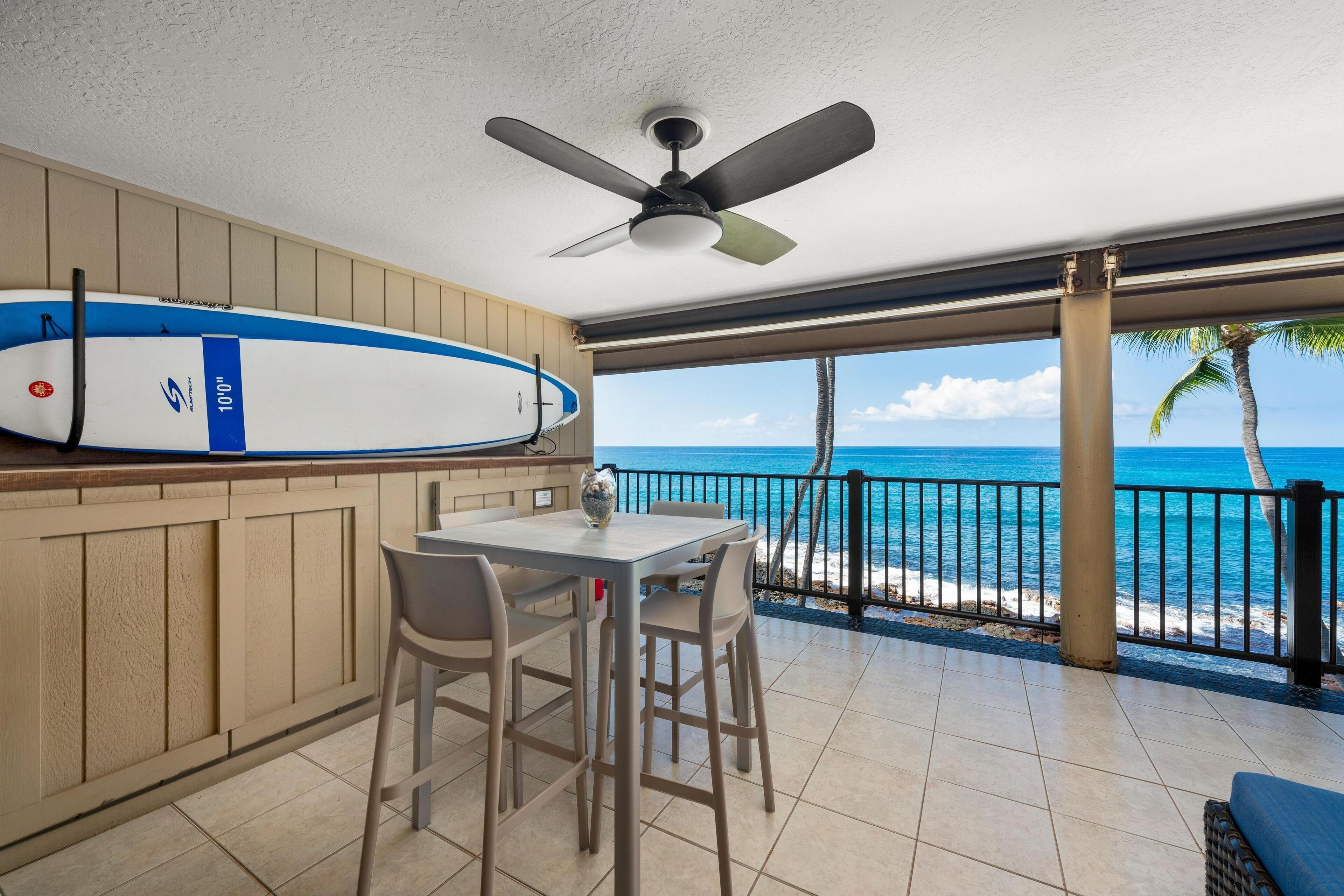 Un balcon avec vue sur l’océan, une planche de surf et un ventilateur de plafond.