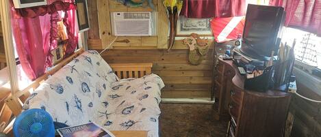 Un interior de autocaravana acogedor con suelo de madera, un sofá, una pequeña mesa con un ventilador, un televisor y una estantería con libros y objetos decorativos.