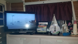 Un téléviseur posé sur un meuble, avec un lecteur DVD, une maquette de petit bateau et une maquette de phare sur une commode.