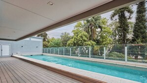 En swimmingpool med træterrasse, omgivet af et glasgelænder og træer.