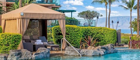 A pool area with lounge chairs and a tent, surrounded by tropical plants and a view of the ocean.