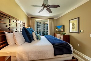 Ein Hotelzimmer mit einem großen Bett, einem Deckenventilator, Blick auf ein Gebäude durch ein Fenster und einem gerahmten Bild an der Wand.
