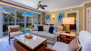 A modern living room with a sofa, armchairs, a coffee table, and a ceiling fan. Large windows offer a view of a pool and outdoor area. - Honua Kai Hokulani by Coldwell Banker Island Vacations (Lahaina)