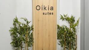 A wooden sign with the text "Oikia suites" in a hallway with white walls, two bamboo plants, and a stone pathway.