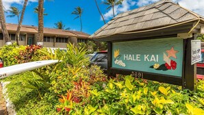 A sign for Hale Kai with a tropical garden and palm trees in the background. - Hale Kai by Coldwell Banker Island Vacations (Lahaina)