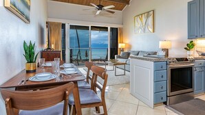 A modern kitchen with a built-in oven, a dining area with a table set for four, and a view of the ocean through large windows. - Hale Kai by Coldwell Banker Island Vacations (Lahaina)