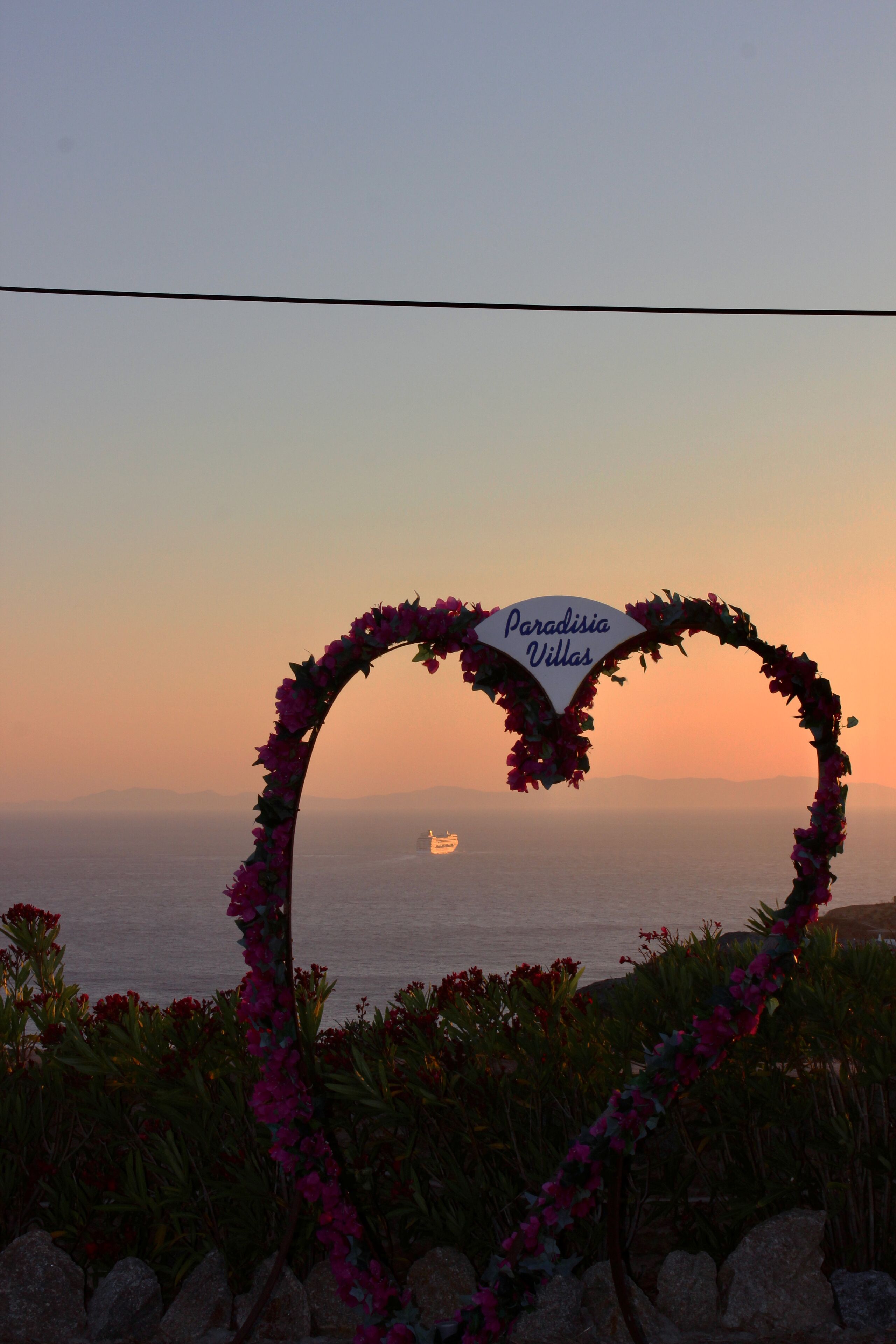 Une arche florale en forme de cœur, sur laquelle est inscrit le nom « Paradisia Villas », se dressant sur fond de coucher de soleil.