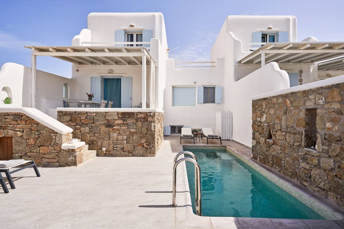A pool area with stone walls and white buildings, including a balcony with a table and chairs.