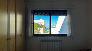 Een slaapkamer met een bed, een raam met uitzicht op gebouwen en de zee, een radiator en een kledingkast.