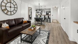 Een moderne woonkamer met een bruine leren bank, een salontafel en een grote wandklok. Er is een plafondventilator en een keuken in de achtergrond.