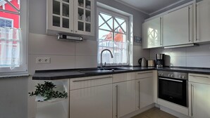 A modern kitchen with white cabinets, a stainless steel refrigerator, a built-in oven, and a window with a view of a red building. - Doppelhaushälfte für 6 Personen mit Wlan (Graal-Müritz)