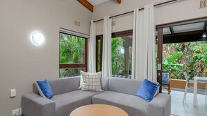 A modern living room with a grey sectional sofa, a round wooden coffee table, and large windows offering a view of lush greenery. - San Lameer Villa 3204 (Southbroom)