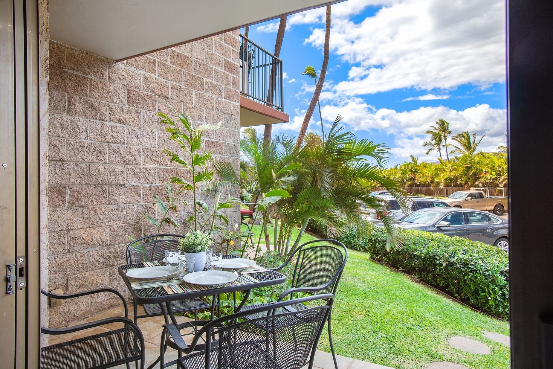 An outdoor patio with a brick wall, a table set for dining, and a view of parked cars and palm trees.
