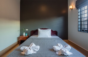 Ein Hotelzimmer mit kariertem Bettbezug, einem Nachttisch mit Vase und Lampe sowie einem Fenster mit Jalousien.