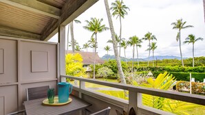 A balcony with a table and chairs, overlooking a garden with palm trees and a view of distant mountains. - Kauai Plantation Hale Suites by Coldwell Banker Island Vacations (Kapaa)