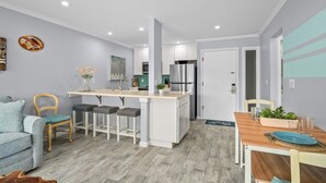 A modern kitchen with white cabinets, a central island, and a dining area with a wooden table and chairs. - Kauai Plantation Hale Suites by Coldwell Banker Island Vacations (Kapaa)