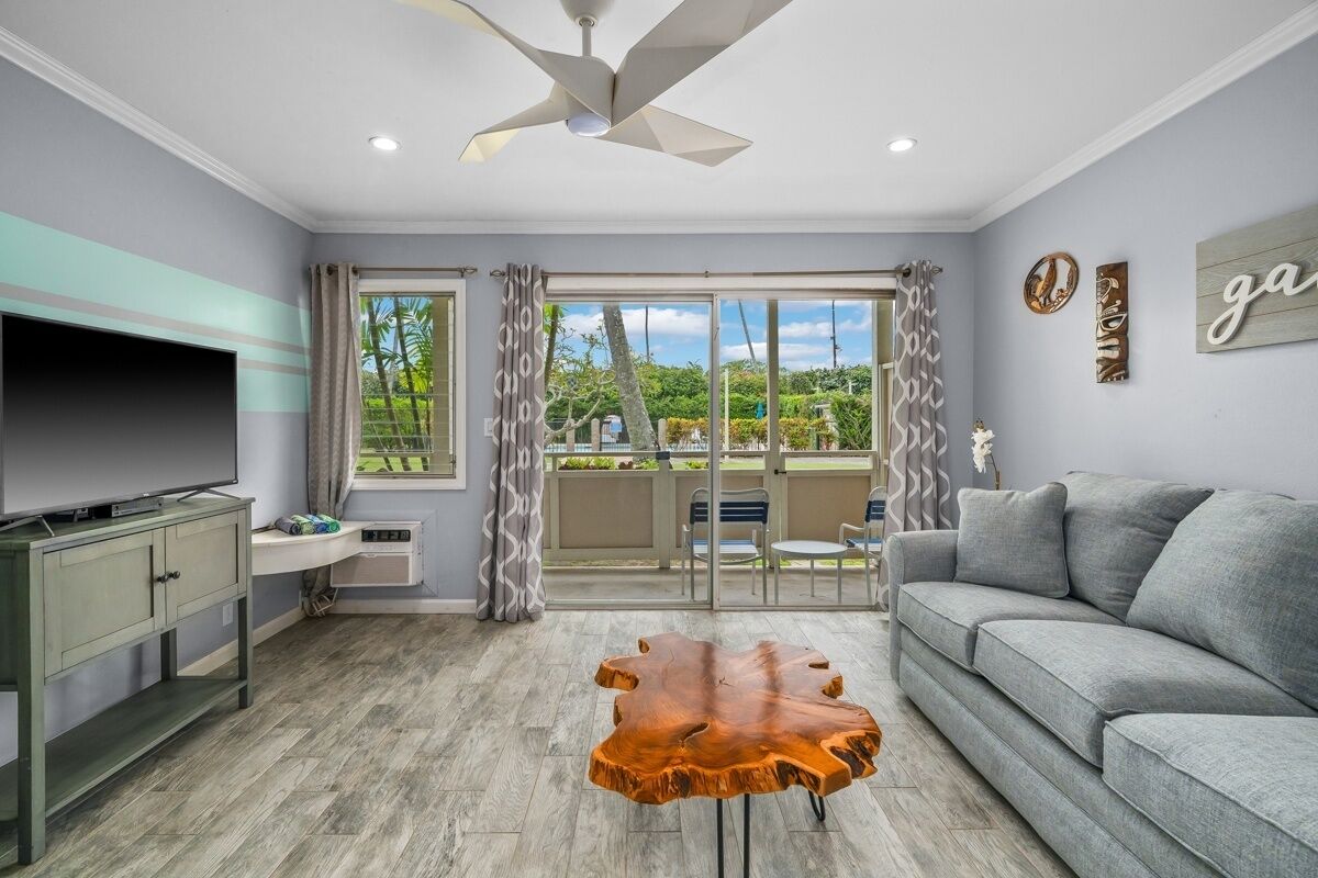 A living room with a flat-screen TV, a grey sofa, a wooden coffee table, and a ceiling fan.