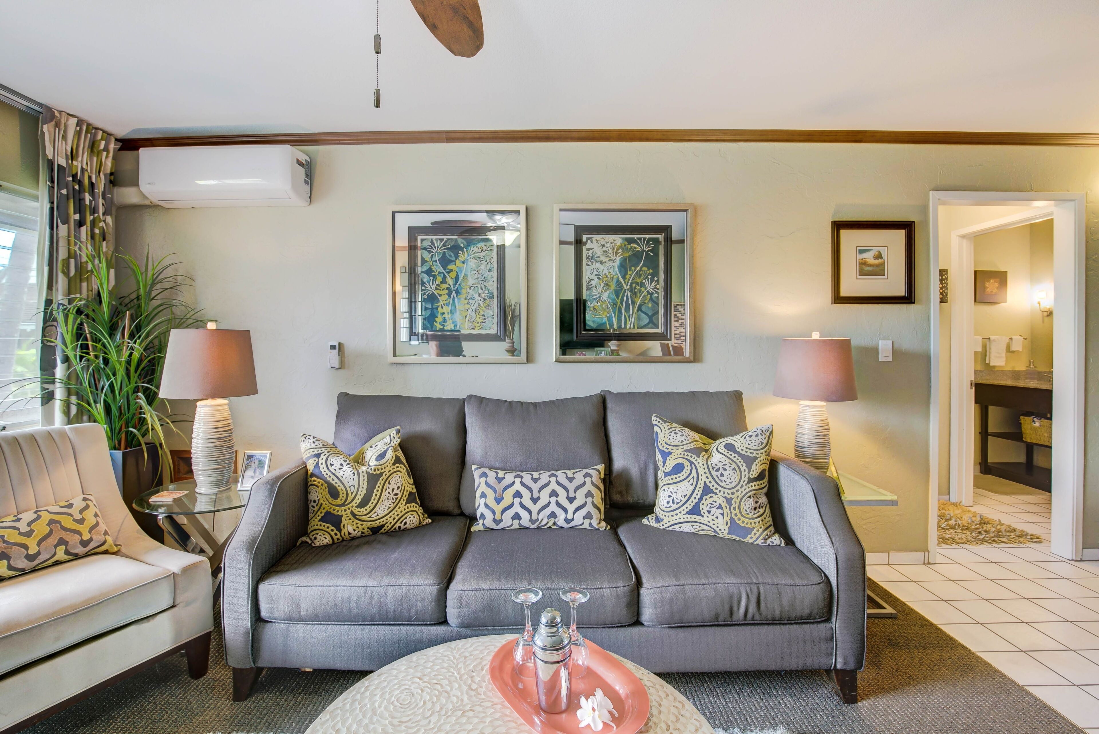 A living room with a grey sofa, patterned cushions, a glass coffee table, and two wall-mounted air conditioners.