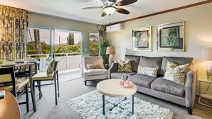 A modern living room with a grey sofa, a round coffee table, and a ceiling fan. There are two framed artworks on the wall, a window with a view of greenery, and a dining area with chairs and a table. - Kauai Plantation Hale Suites by Coldwell Banker Island Vacations (Kapaa)