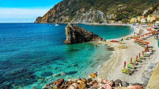 A coastal beach with clear turquoise waters, a rocky outcrop, and colorful umbrellas along the shore.
