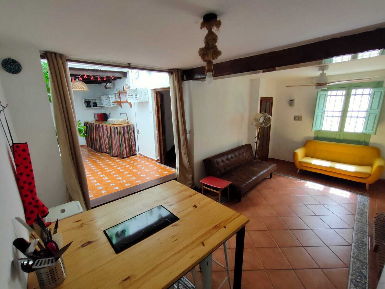 Una sala de estar con un sofá marrón, un sofá amarillo, una mesa de madera y una cocina visible a través de una puerta abierta.