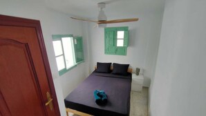 Une pièce avec un lit, un ventilateur de plafond, une fenêtre avec des volets verts et une porte en bois.