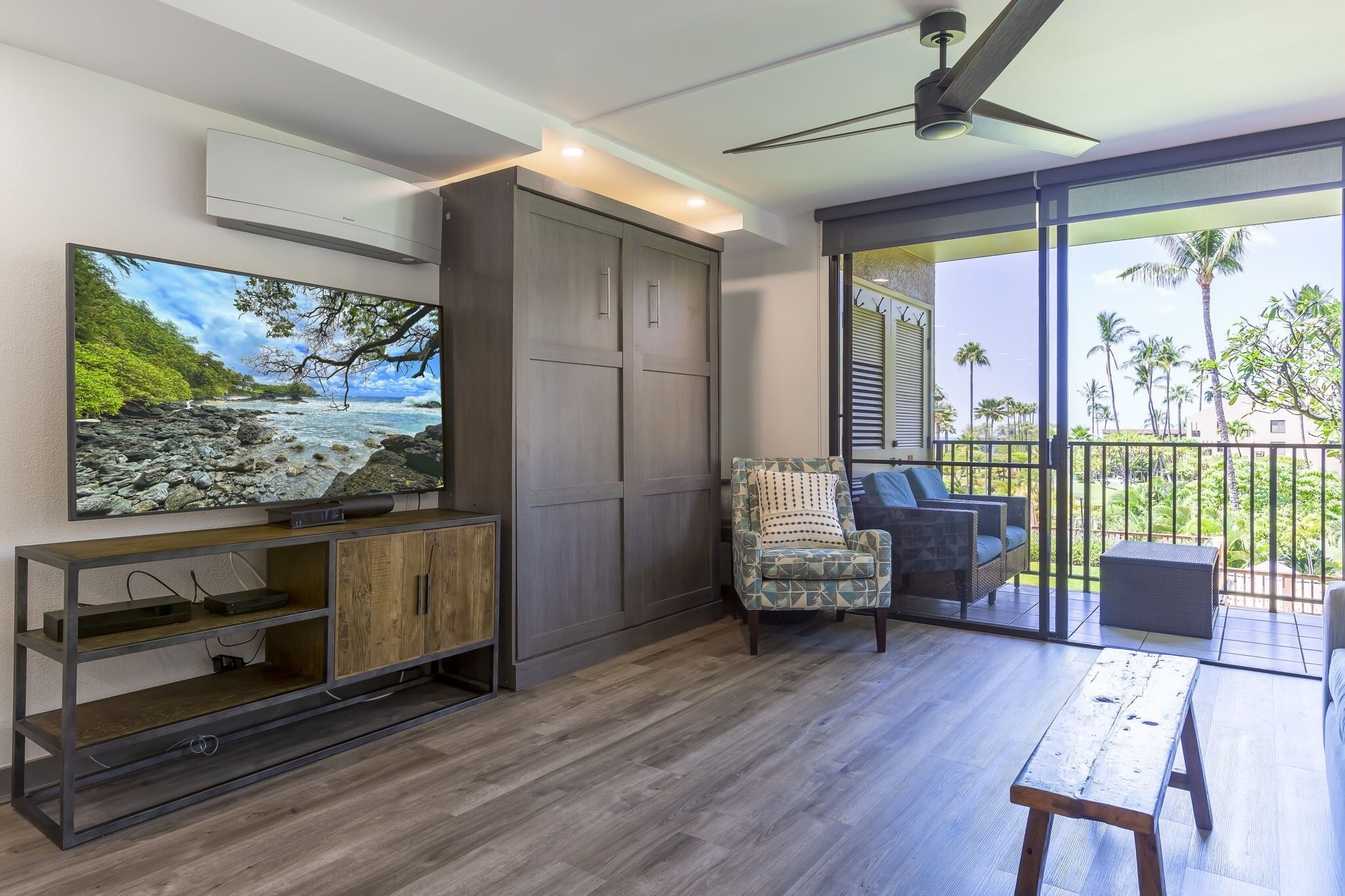 Una sala de estar moderna con un televisor de pantalla plana grande, piso de madera, un armario gris, un sillón estampado y un balcón con vista a palmeras.