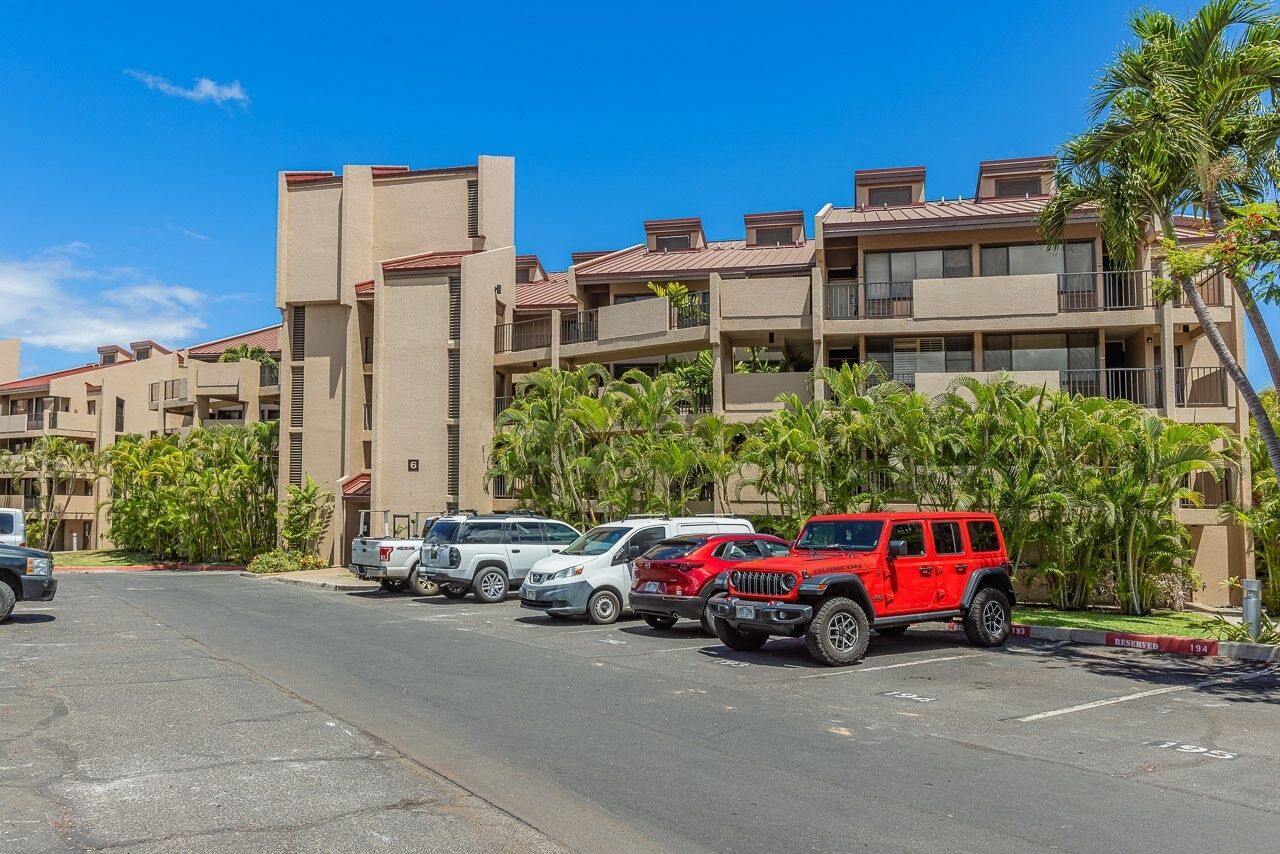 A parking lot with several cars, including a red Jeep, in front of a building with balconies and palm trees.