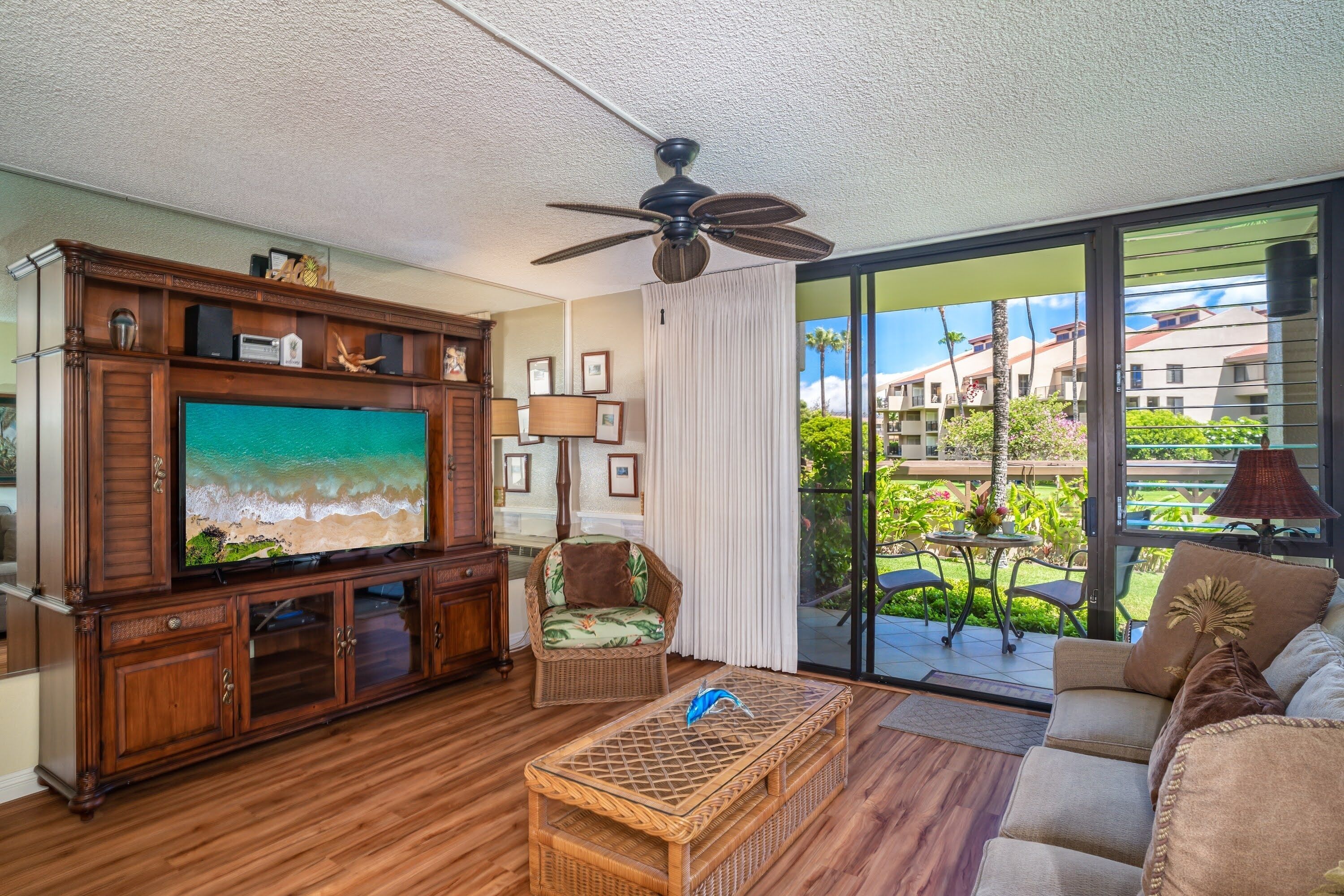 A spacious living room with a large flat-screen TV, a ceiling fan, and a view of a balcony with outdoor furniture.