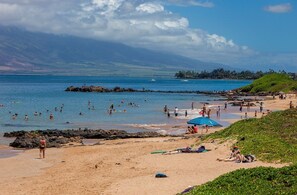 Una playa con gente nadando y tomando el sol, una zona rocosa y vistas a montañas distantes.