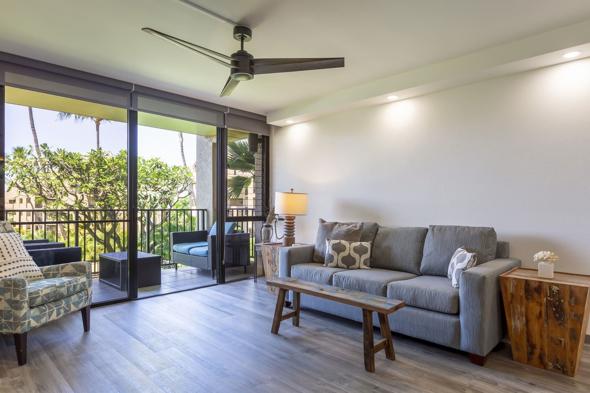 A spacious living area with a grey sofa, a wooden coffee table, and a ceiling fan. A balcony with a glass door is visible, offering a view of palm trees and a building.