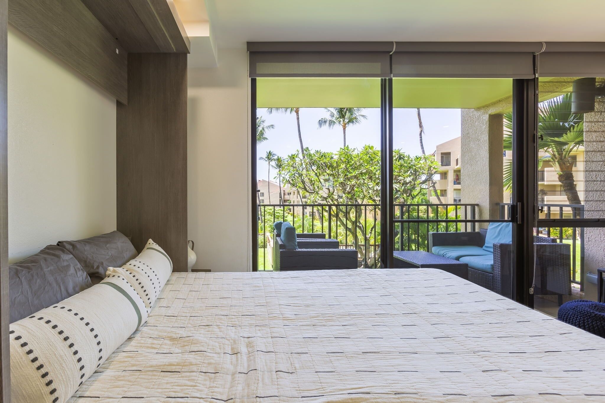 Une chambre d’hôtel avec un grand lit, un balcon doté d’un espace salon extérieur et une vue sur les palmiers et les immeubles.