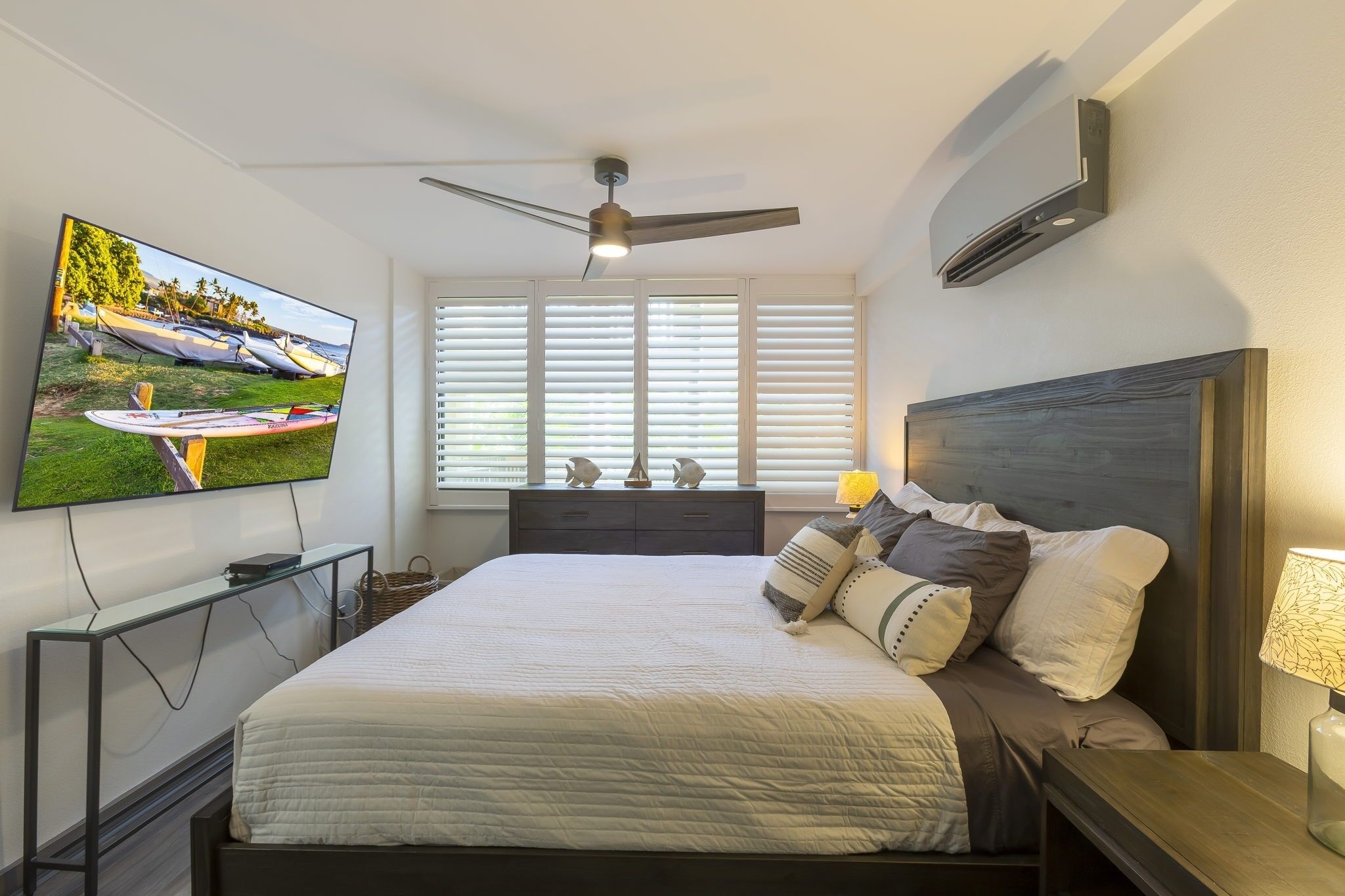 Un dormitorio con una cama, un televisor montado en la pared, un ventilador de techo y una ventana con persianas.