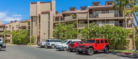 A parking lot with several cars, including a red Jeep, in front of a building with balconies and palm trees.