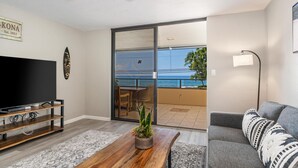 A living room with a flat-screen TV, wooden coffee table, grey sofa, and a balcony with a view of the ocean. - Big Island Holualoa Bay Villas by Coldwell Banker Island Vacations (Kailua-Kona)