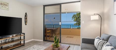 A living room with a flat-screen TV, wooden coffee table, grey sofa, and a balcony with a view of the ocean.