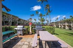 A patio area with two barbecue grills, picnic tables, and a view of a hotel building with palm trees.