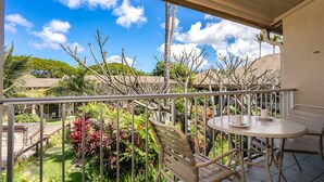 A balcony with a white table and chairs, surrounded by tropical plants and a clear blue sky. - Napili Shores by Coldwell Banker Island Vacations (Lahaina)