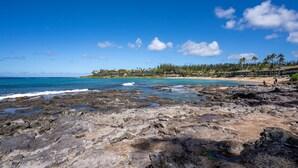 A rocky shoreline with a clear blue sky, a sandy beach, and a line of buildings in the background. - Napili Shores by Coldwell Banker Island Vacations (Lahaina)
