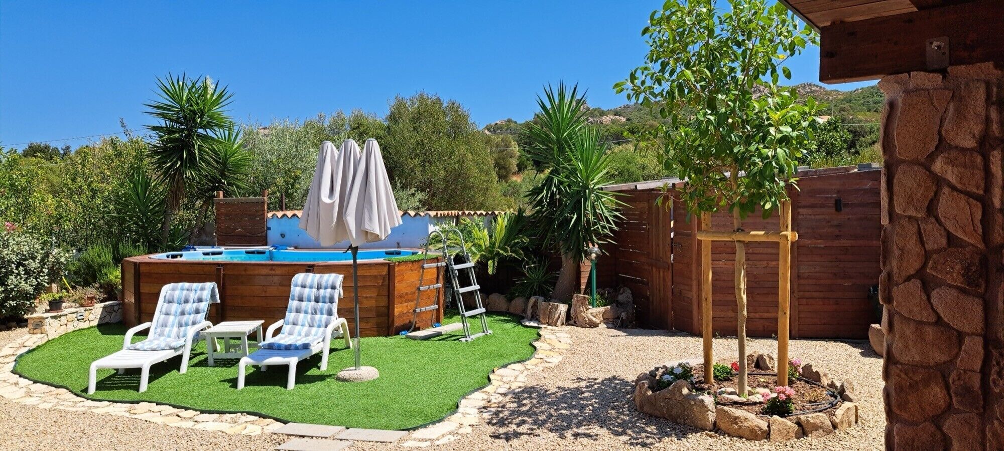 Un patio trasero con un jacuzzi, dos sillones reclinables, una terraza de madera y un árbol pequeño.