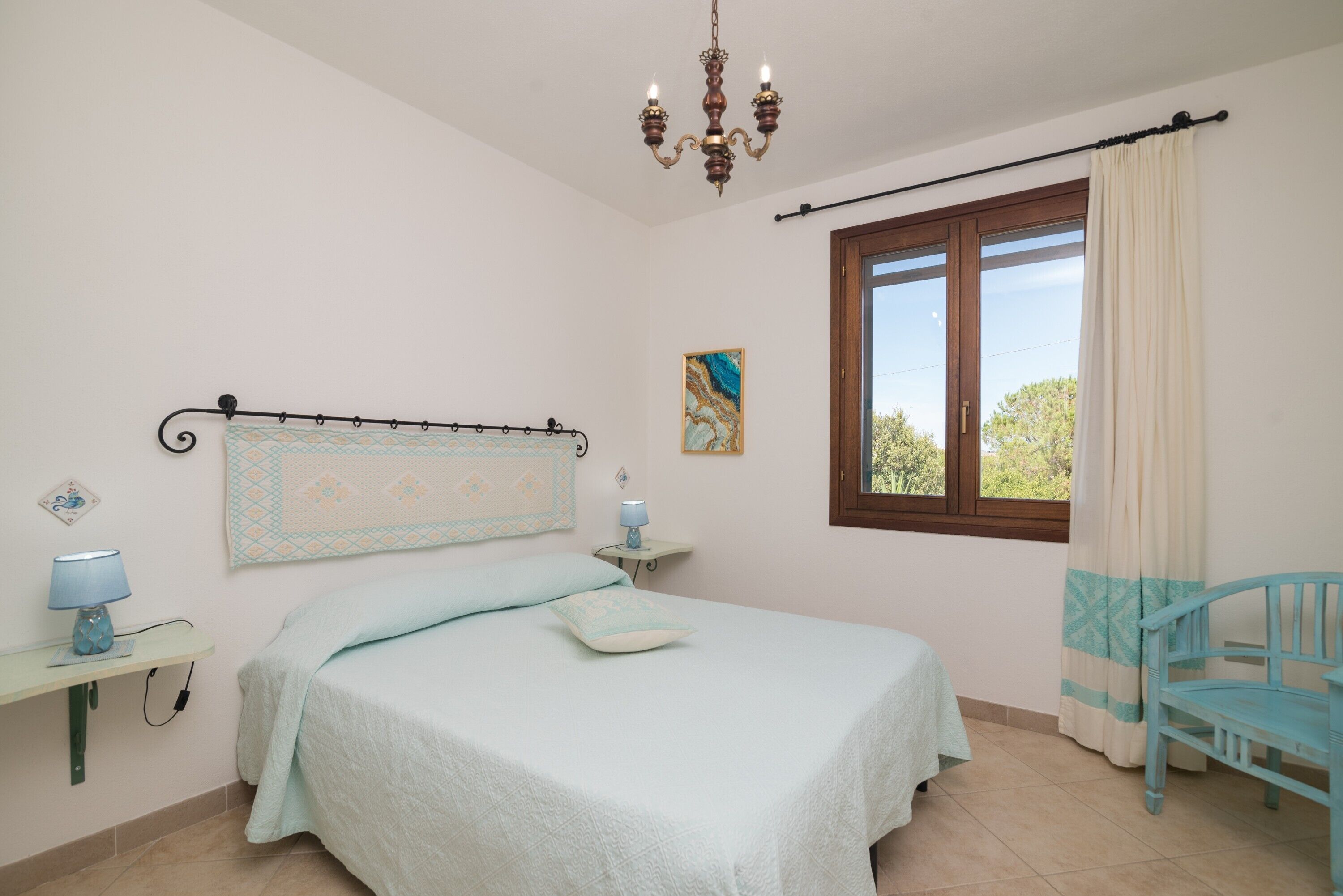 Ein Schlafzimmer mit Bett, Nachttisch, Lampe, Stuhl, Fenster und einer Chandelier.