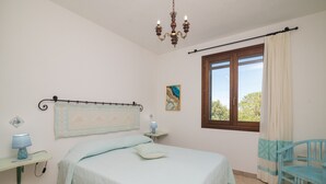 A bedroom with a bed, bedside table, lamp, chair, window, and chandelier.