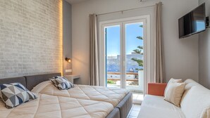 A hotel room with a brick wall, a bed with a quilted mattress, a sofa, a flat-screen TV, and a balcony with a view. - Holiday Home 'Porto Galini Tinos Perastra 103' with Sea View, Wi-Fi and Air Conditioning (?a??t?)