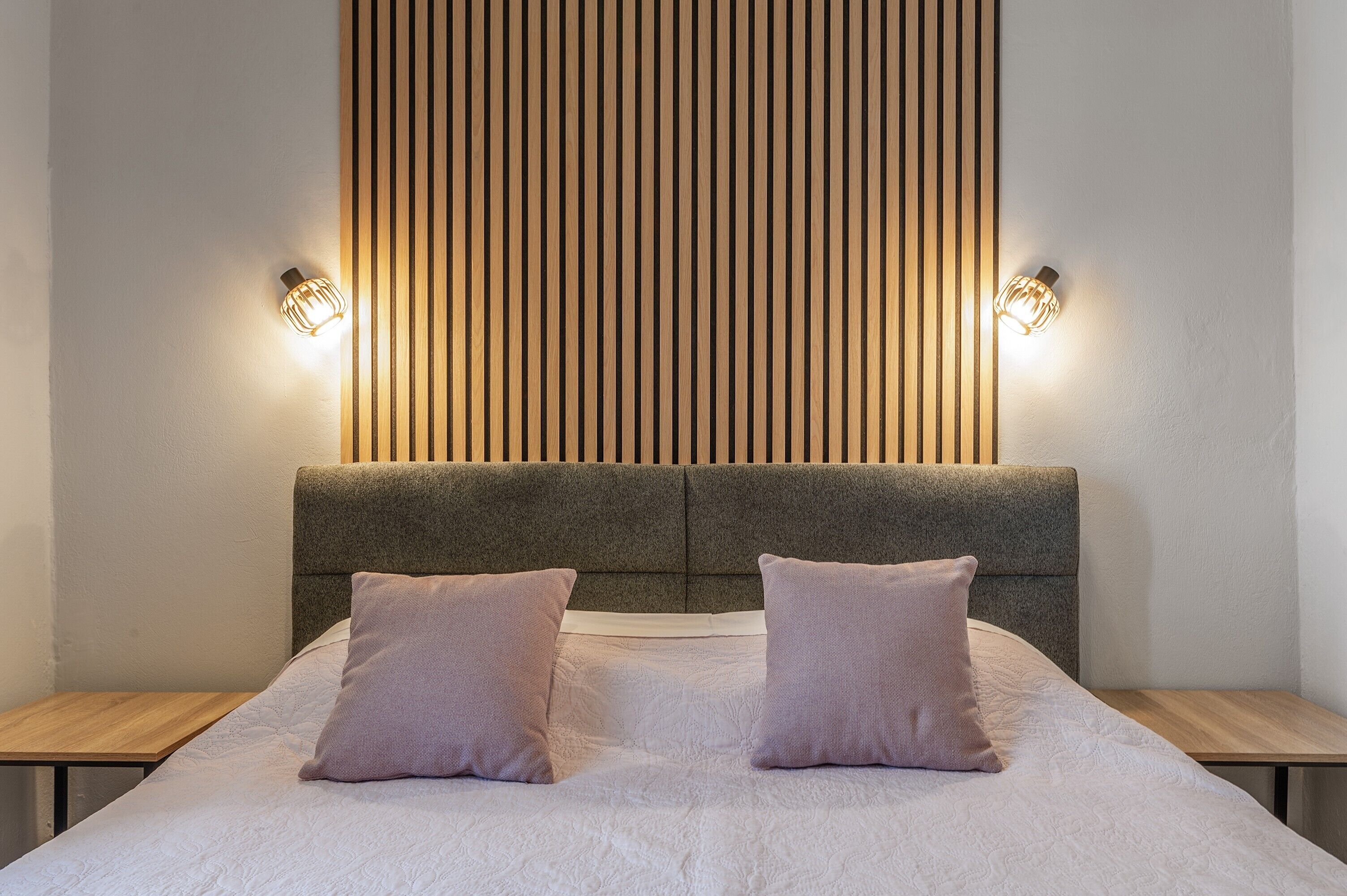 Una cama con cabecera gris, dos mesitas de noche e iluminación empotrada en la pared.