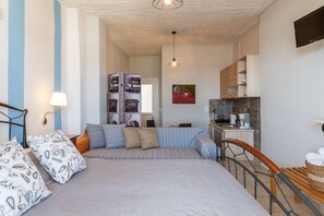 Quarto com cama, travesseiros, mesa de cabeceira, luminária, sofá, cozinha compacta com armários, micro-ondas e televisão.