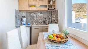 A compact kitchen with wooden cabinets, a tiled backsplash, a dining table with a fruit bowl, and a window with a view of buildings and hills. - Holiday Home 'Porto Galini Tinos Myrsini 106' with Private Terrace, Wi-Fi and Air Conditioning (LAOUTI)