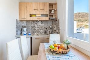 A compact kitchen with wooden cabinets, a tiled backsplash, a dining table with a fruit bowl, and a window with a view of buildings and hills.